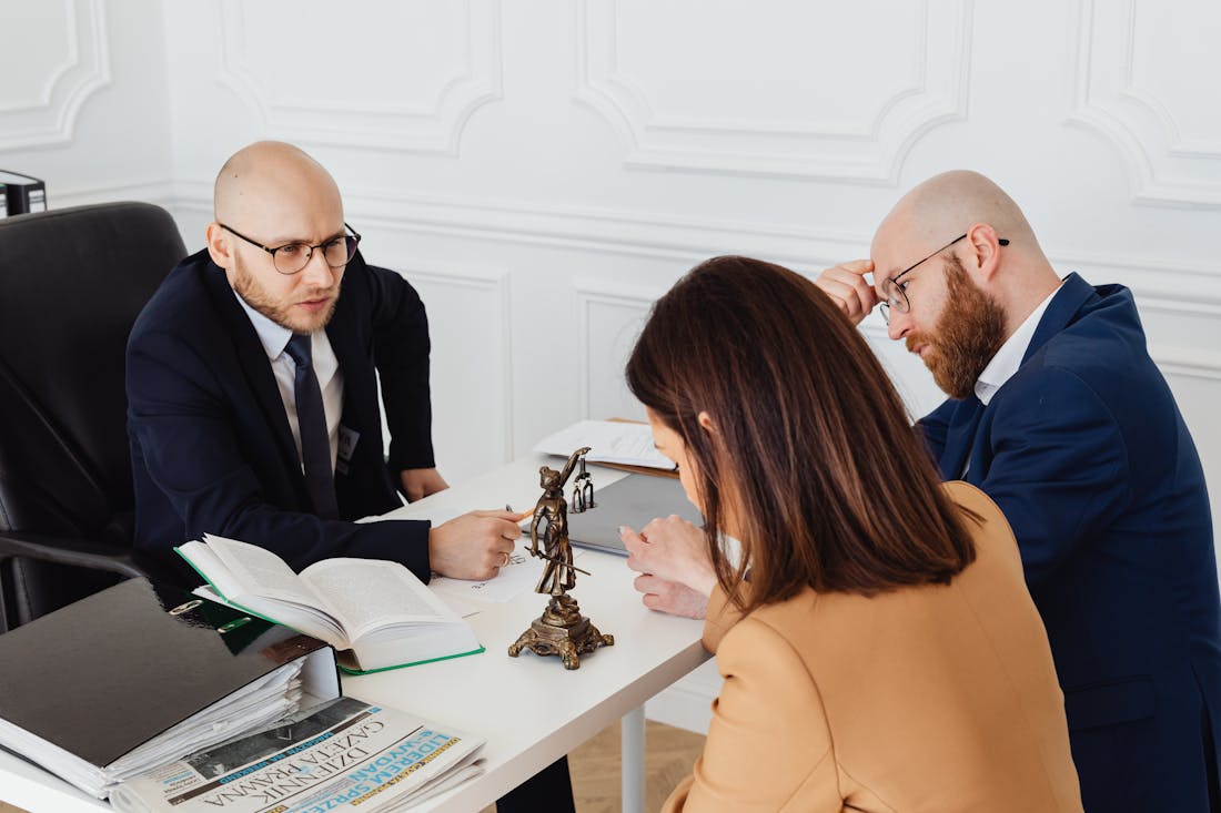 An attorney meeting with two clients across a desk with law books and a scales-of-justice statue